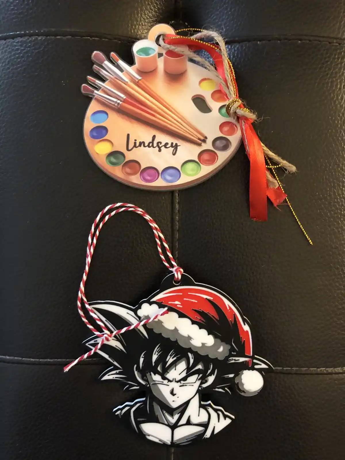 Two Christmas ornaments rest on a black leather surface. The top one is a circular paint palette design with brushes and…