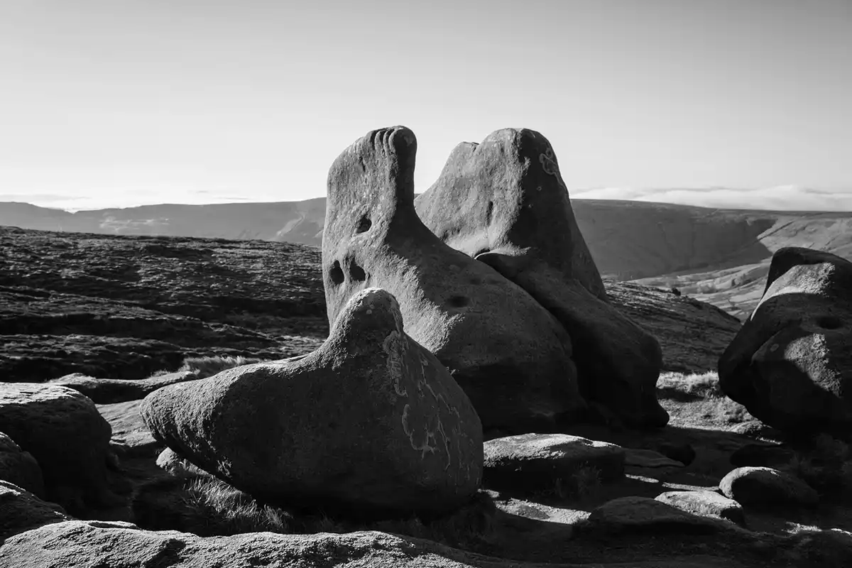 A black-and-white photograph of large, oddly shaped boulders in a barren, hilly landscape. The central boulders are arranged…